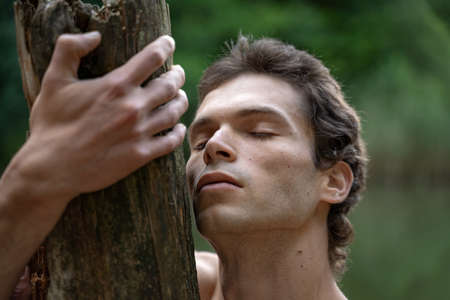 Close-up of shirtless man with eyes closed embracing wood in forestの写真素材