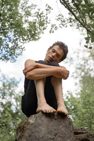 Low angle view of man with eyes closed hugging knees while sitting on rock in woodlandの写真素材