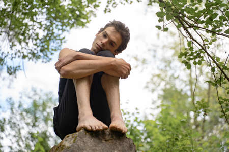 Low angle portrait of man hugging knees while sitting on rock in forestの写真素材