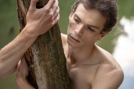 Close-up of thoughtful shirtless man embracing wood looking away in forestの写真素材