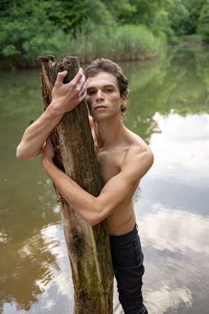 Portrait of shirtless man hugging wood while standing against lake in forestの写真素材