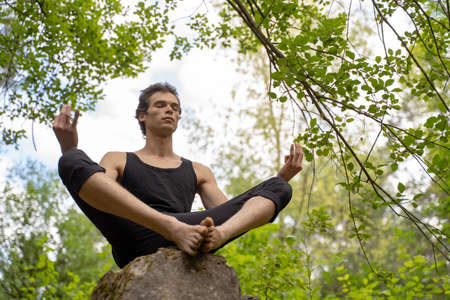 Low angle view of man with eyes closed meditating while sitting on rock in forestの写真素材