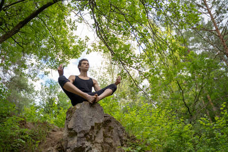 Low angle view of man meditating while sitting on rock against trees in forestの写真素材