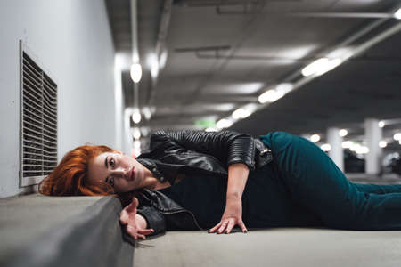 Attractive millennial girl with head on curb laying on floor in parking house. Female model with red hairs and open eyes. Acting dead victim of crime.の写真素材
