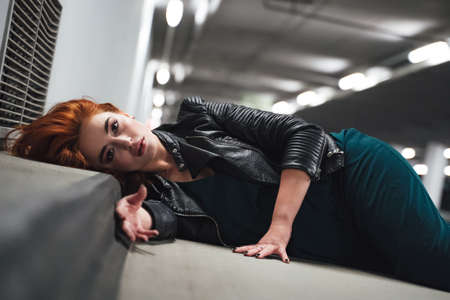 Closeup of beautiful millennial girl laying on floor in parking house with head on curb. Female model with red hairs, open eyes. Acting dead victim.の写真素材