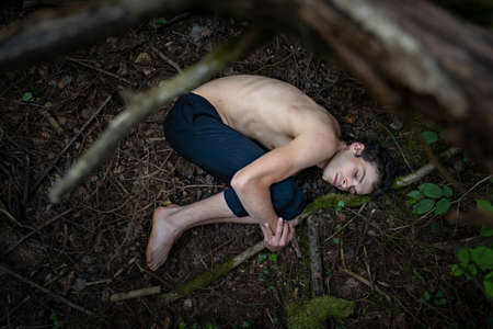 High angle view of shirtless man hugging knees while sleeping on dirty land in forestの写真素材
