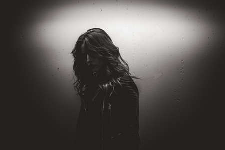Black and white low contract portrait of female model. Long-haired girl in leather jacket standing at white spot illuminated wall.の写真素材