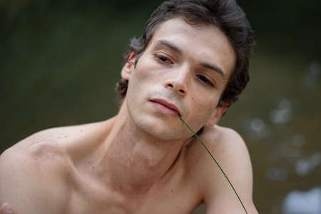 Close-up of shirtless serious man with plant stem in mouth against lake at forestの写真素材