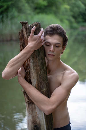 Shirtless man with eyes closed embracing wood while standing against lake in forestの写真素材