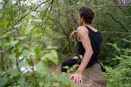 Rear view of man sitting on rock amidst trees in forestの写真素材
