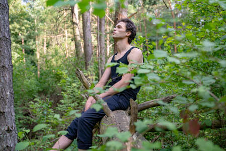 Side view of thoughtful man looking away while sitting on log against trees in forestの写真素材