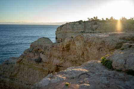 Distant view of couple standing on cliff by sea against clear sky at sunsetの写真素材