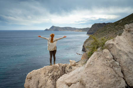 Rear view of woman with arms outstretched looking at sea against sky while standing on rockの写真素材