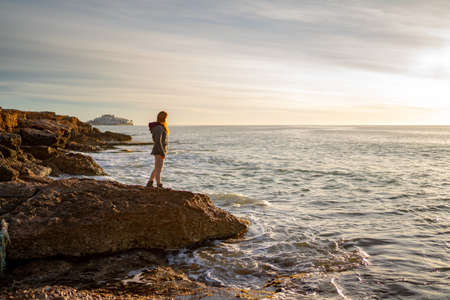 Side view of woman looking at seascape while standing on rock against sky during sunsetの写真素材