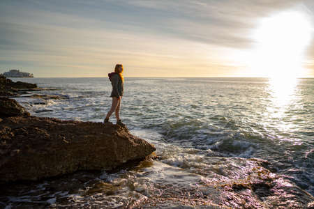Side view of woman looking at seascape against sky while standing on rock during sunsetの写真素材