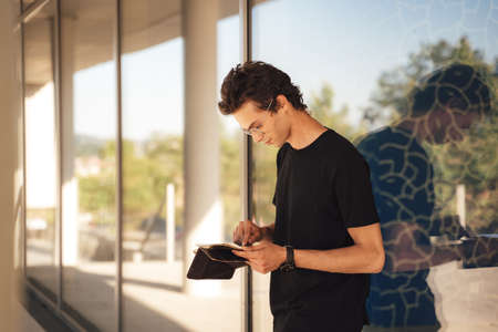 Young man using digital tablet while standing against glass window at college campusの写真素材