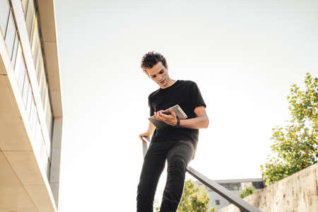 Businessman sitting on railing and using digital tablet outside office against skyの写真素材