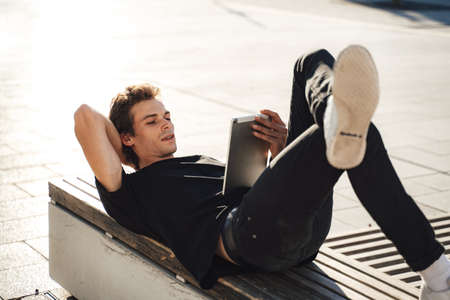 Full length of European young man using digital tablet while lying on bench at college campusの写真素材