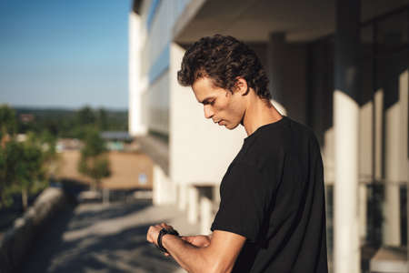 Young man watching time on his wrist watch while standing outside university buildingの写真素材