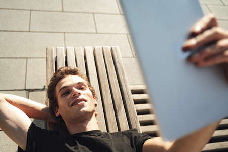 Top view of handsome young man having video call on tablet computer while lying on benchの写真素材