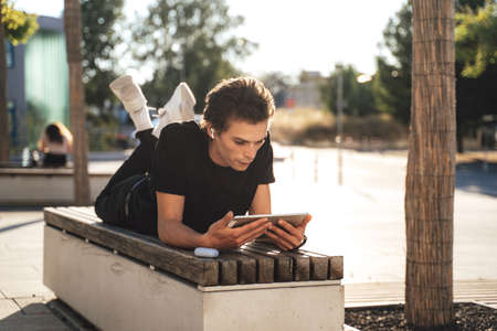 Young man watching movie on digital tablet while lying on bench at college campusの写真素材