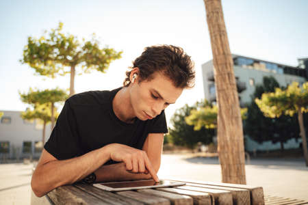 Young male university student touching screen of digital tablet while lying on bench at campusの写真素材