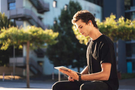 Handsome young man studying online on digital tablet while sitting at college campusの写真素材