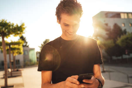 Smiling young man reading text message on smartphone in city on sunny dayの写真素材