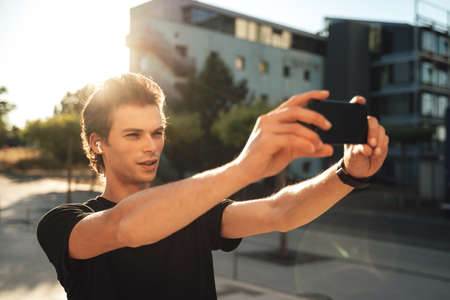 Caucasian young man photographing using smartphone in city on sunny dayの写真素材