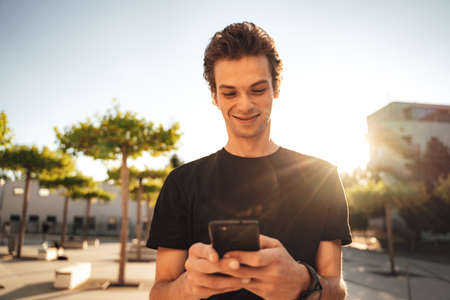 Handsome Caucasian university student smiling while using smartphone in city on sunny dayの写真素材