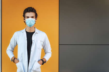 Portrait of scientist or doctor wearing mask and white coat with hands in pockets standing against...