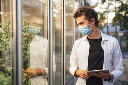 Young male scientist holding digital tablet while examining plants in glasshouse during COVID-19 outbreakの写真素材
