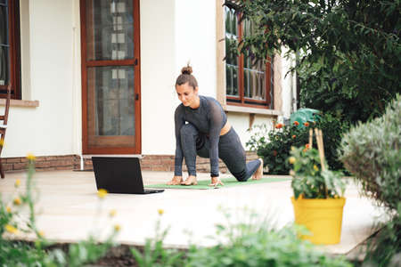 Fit young woman learning stretching exercise while watching video on laptop in backyardの写真素材