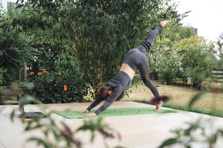 Active young woman in three legged downward dog pose on exercise mat with laptop in backyardの写真素材