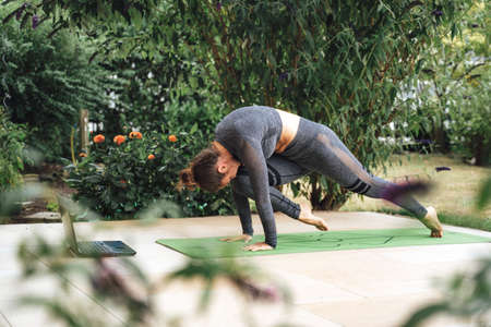 Full length of sporty Caucasian woman practicing tiger curl pose on yoga mat with laptop at patioの写真素材