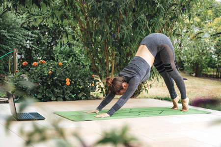 Caucasian woman in sportswear practices yoga downward facing dog pose with laptop in backyardの写真素材