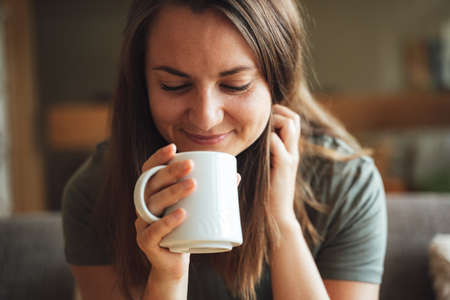 Close up of attractive brunette Caucasian woman enjoying coffee from mug at homeの写真素材