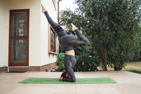 Full length side view of active young woman practicing headstand exercise on yoga mat at patioの写真素材