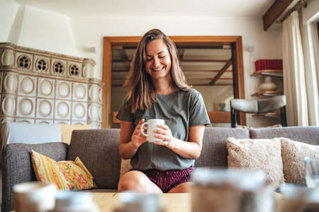 Beautiful young woman smiling while having morning coffee sitting on sofa in living room at homeの写真素材