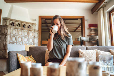 Caucasian young woman drinking coffee while sitting on sofa in living room at homeの写真素材