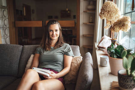 Portrait of attractive young woman holding book while relaxing on couch in living room at homeの写真素材