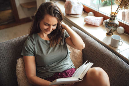 High angle view of lovely young woman with brown hair reading book on sofa at homeの写真素材