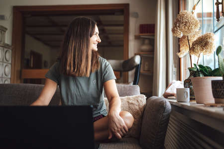 Smiling young woman with brown hair sitting with laptop on sofa at homeの写真素材