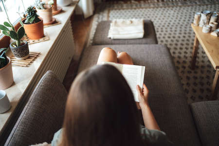Rear view of brunette young woman reading book on couch in living room at homeの写真素材