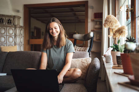 Smiling young woman using laptop while sitting on couch in living room at homeの写真素材