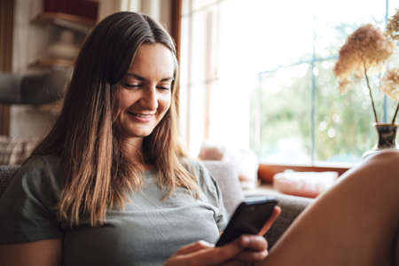 Smiling young brunette woman using smartphone in living room at homeの写真素材