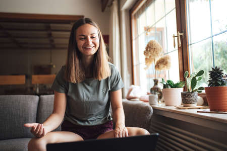 Cheerful young Caucasian woman having a video call on laptop in living room at homeの写真素材