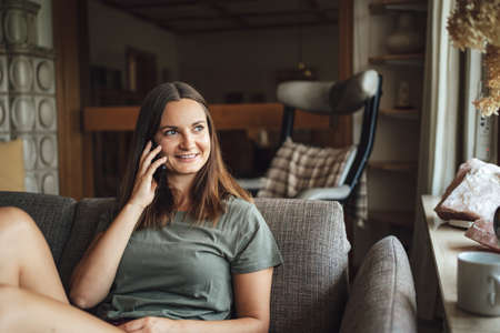 Young Caucasian woman looking away while talking on mobile phone, sitting on sofa at homeの写真素材