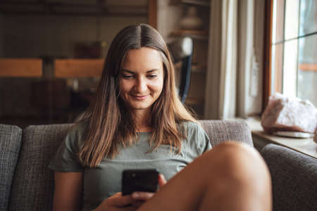 Young brunette in casual sitting on sofa at home and searching in smartphoneの写真素材