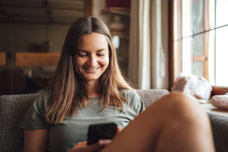 Young brunette woman in casual sitting on sofa at home and using mobile phoneの写真素材